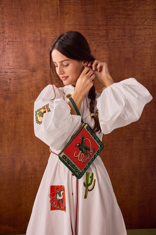 A woman wearing a white long-sleeve dress with colorful embroidered motifs, holding a red and green embroidered crossbody bag, posed against a warm brown textured backdrop.
