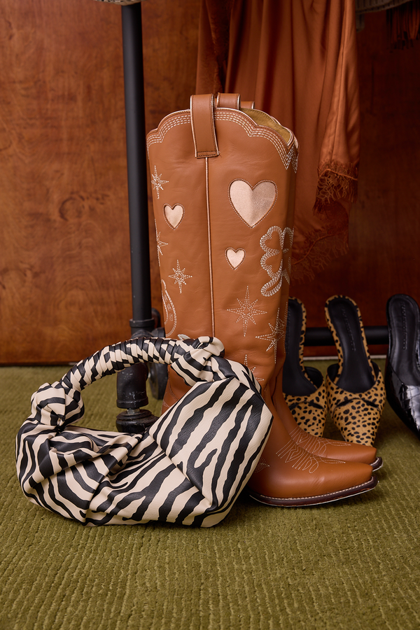 A tan leather cowboy boot with embroidered hearts and floral motifs stands on a green textured rug next to a zebra-print handbag, with additional shoes and warm brown garments in the background.