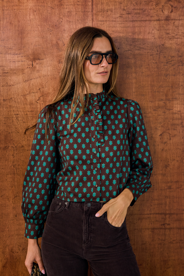 A woman with long light brown hair and black-rimmed glasses wears a dark brown long-sleeve blouse with a teal geometric pattern, paired with dark brown pants, standing against a rustic wooden wall.