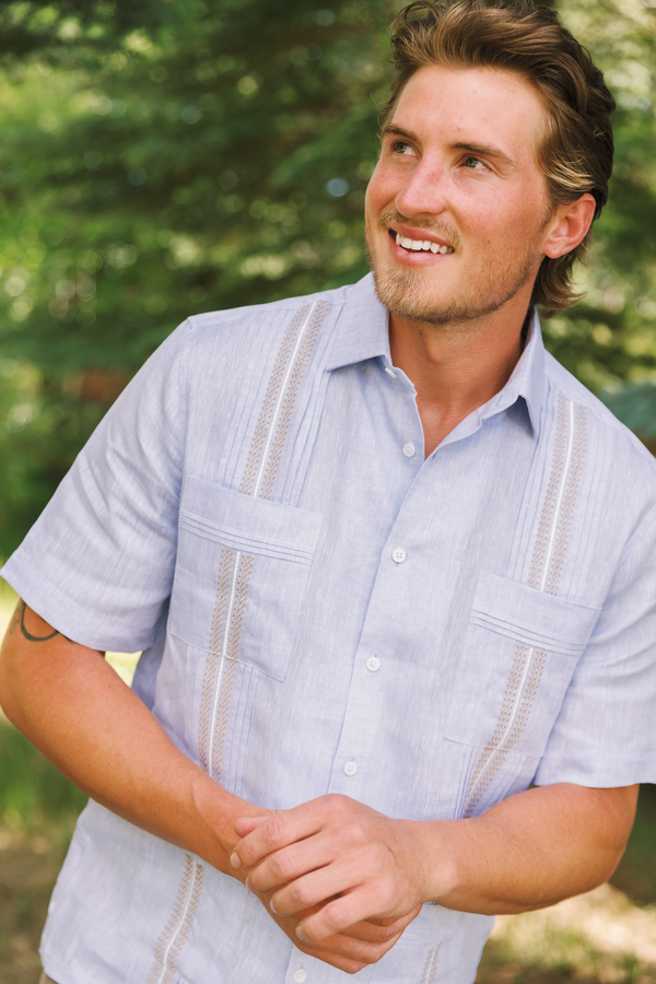 A smiling man wearing a light blue short-sleeve guayabera-style button-up shirt with decorative vertical embroidery, photographed outdoors against a blurred green foliage background.