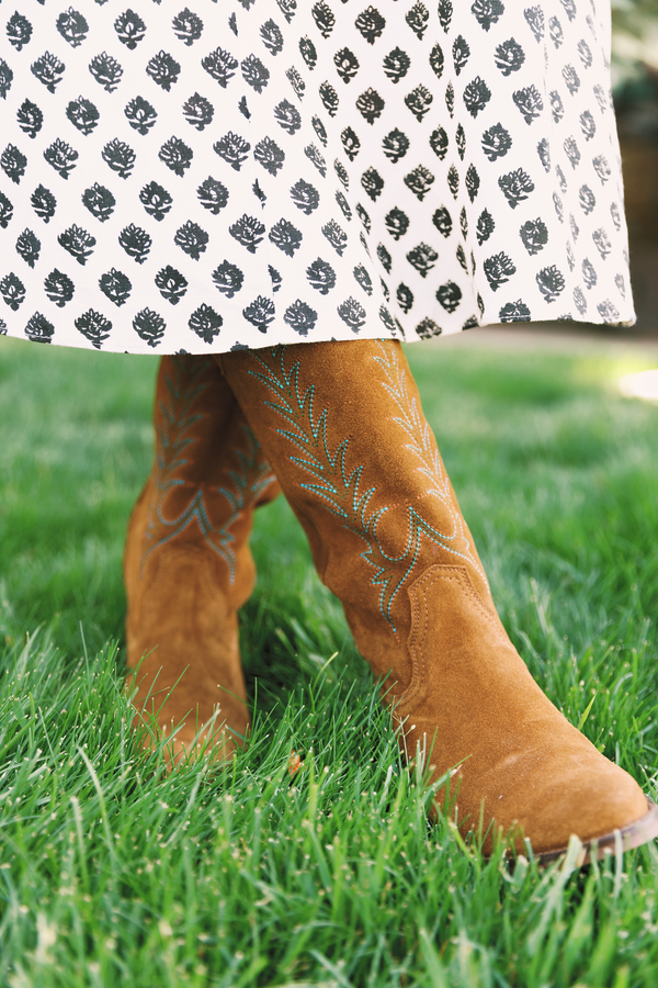 A close-up outdoor shot of a person's legs standing on green grass, wearing tan suede cowboy boots with light blue embroidery and a white skirt with a black leaf pattern.