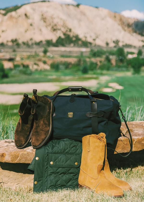 A navy duffel bag with black straps sits outdoors on a stone ledge in a grassy landscape, styled with a pair of dark brown suede boots, a green quilted jacket, and a pair of tan leather boots.