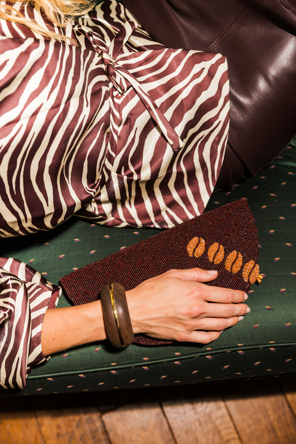 A close-up lifestyle shot of a person wearing a brown and cream zebra-print silk blouse and a dark brown leather skirt, holding a brown clutch and resting on a green patterned sofa.