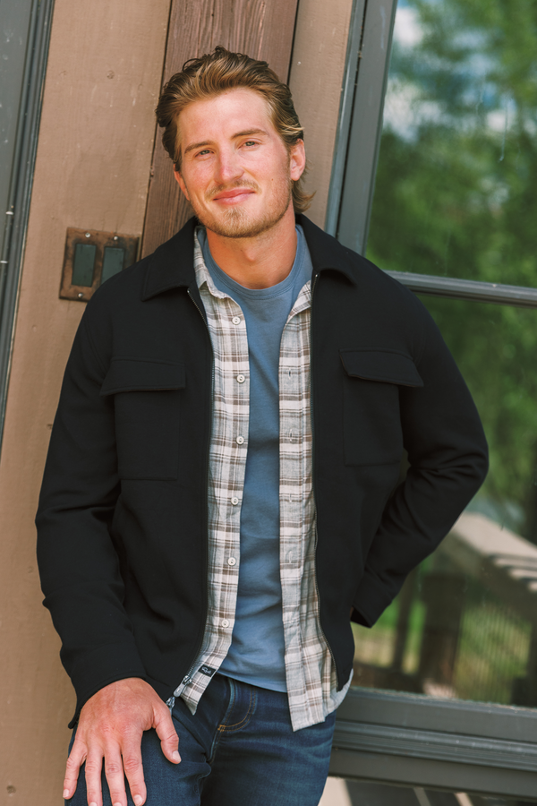 A man wearing a black overshirt over a blue t-shirt and a gray plaid button-down shirt, standing outdoors against a wooden wall and window.