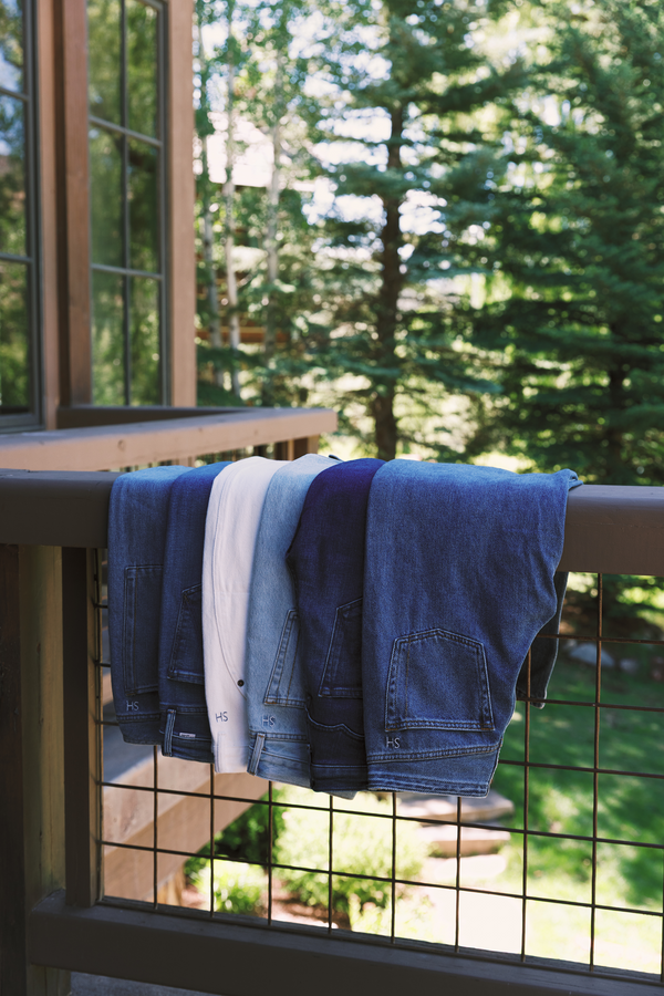 Six pairs of folded jeans in different denim washes hang from a wooden railing on a balcony with a forest background.