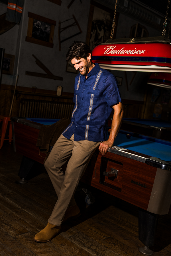 A man wearing a blue short-sleeve Guayabera shirt with white stripes, khaki pants, and tan boots, leaning on a pool table in a dimly lit bar environment.