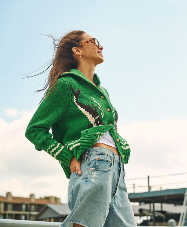 A woman wearing a green knit cardigan with a whale pattern, light-wash denim shorts, and sunglasses, photographed outdoors against a pale blue sky.