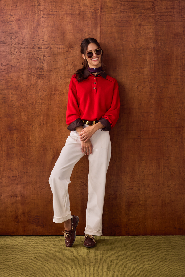 A smiling woman wearing sunglasses, a red button-collar sweater with a dark neck scarf, a gold buckle belt, cream wide-leg trousers, and brown boat shoes, posing against a wood-paneled wall.
