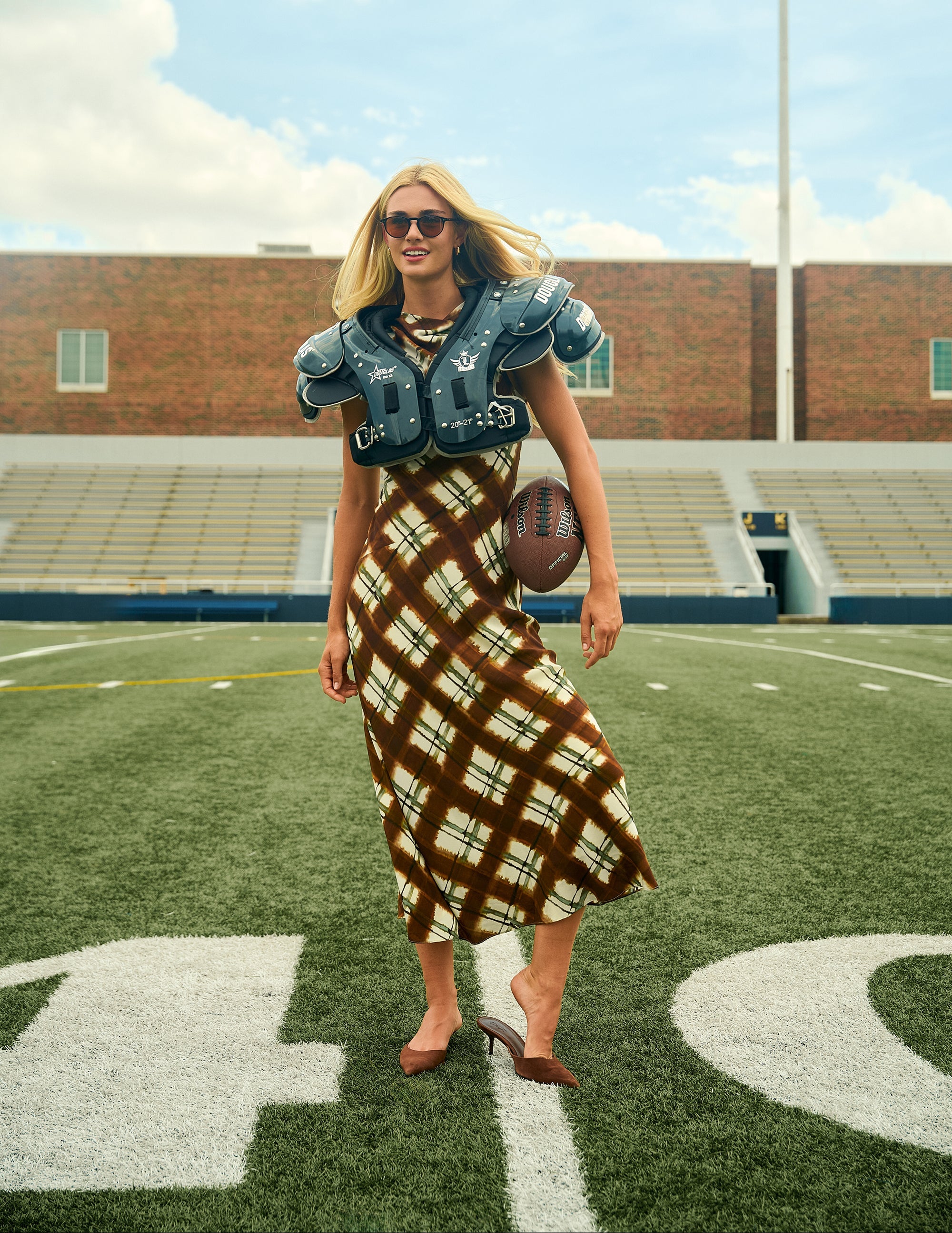 A woman stands on a football field wearing a brown and beige plaid maxi dress, a blue football jersey, and shoulder pads, holding a football. 