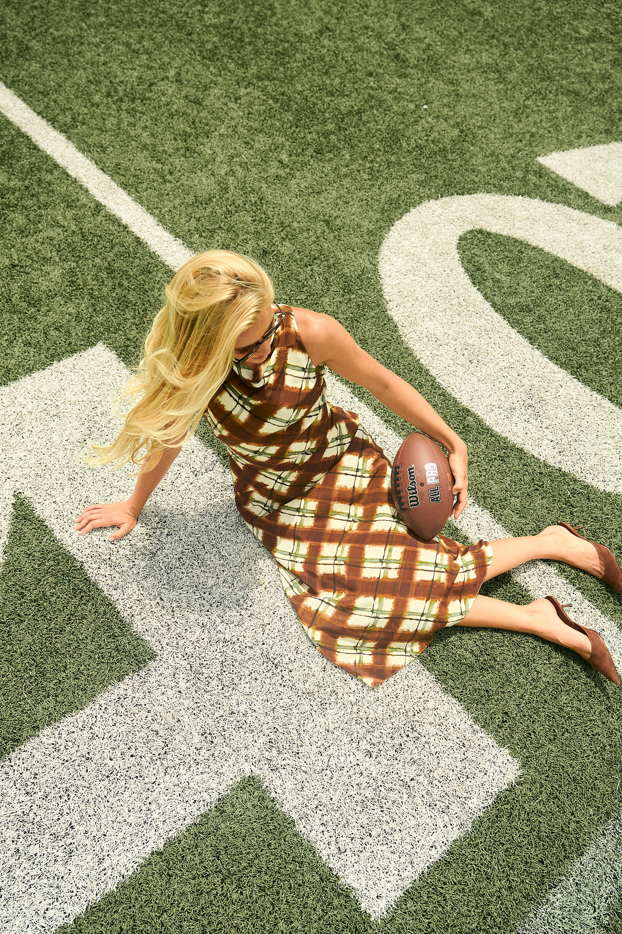A woman in a brown and beige plaid dress sits on a green football field with white yard lines, holding a brown football.