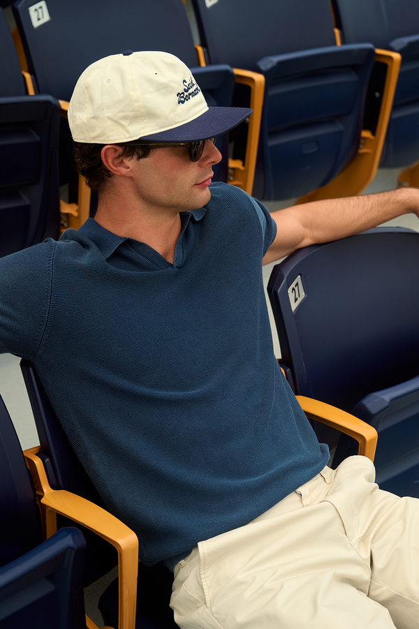 A man wearing a navy blue polo shirt, a cream and navy cap, and sunglasses sits in a row of blue stadium seats.