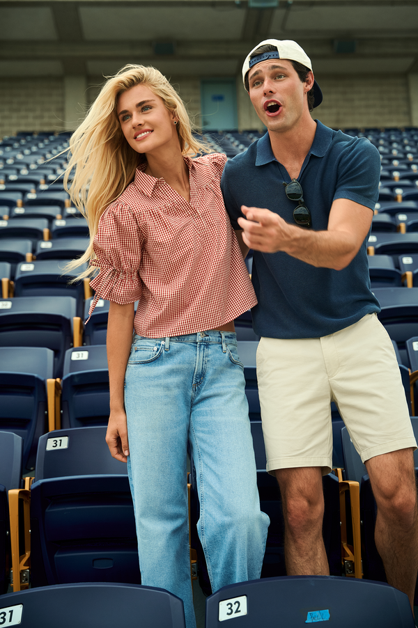 A man and a woman posing together in an indoor stadium with rows of blue seats. The woman wears a pink and white gingham short-sleeve button-up shirt and light blue jeans, while the man wears a navy blue polo shirt, beige shorts, and a white baseball cap.