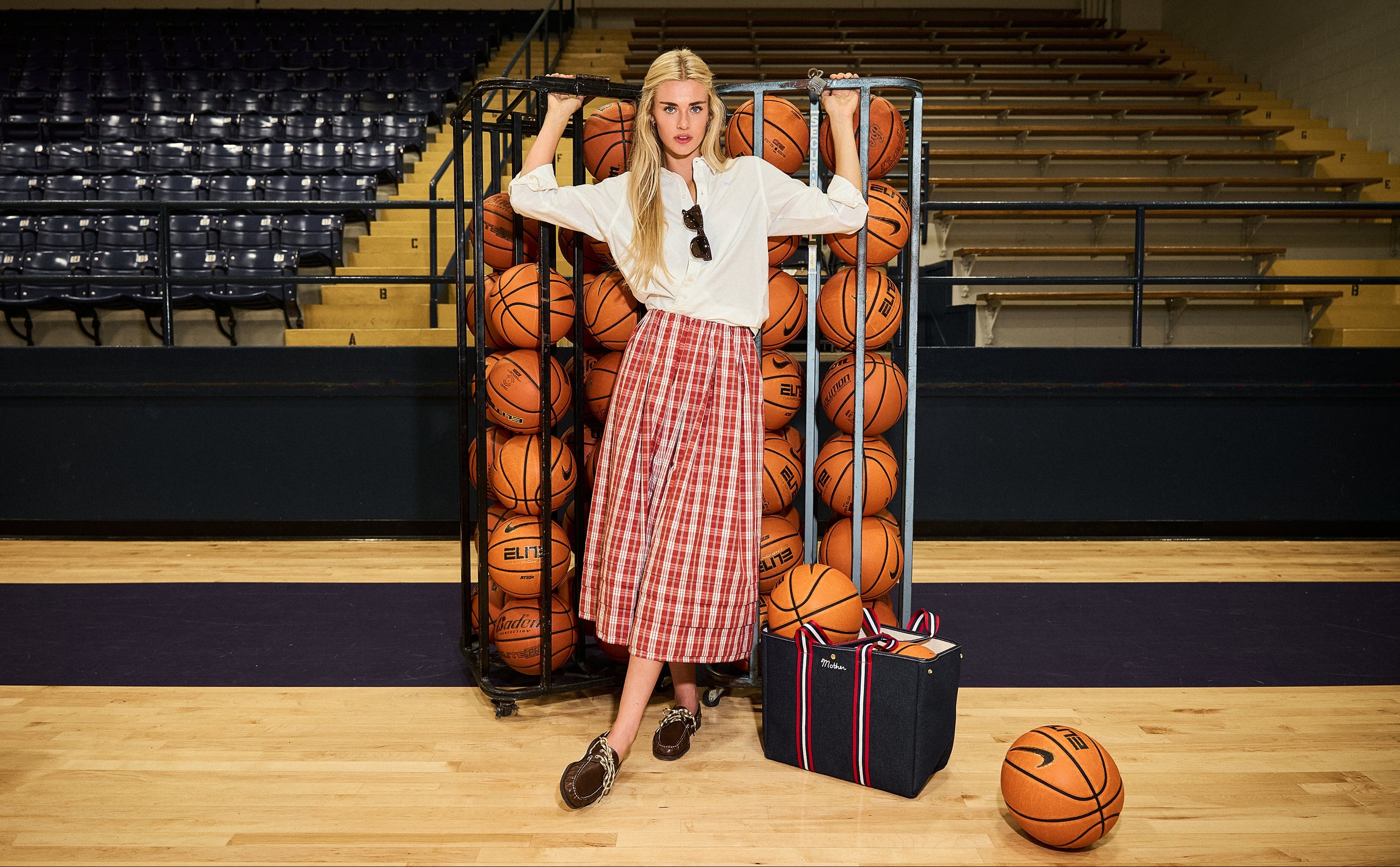 A woman stands on a basketball court in front of a rack filled with basketballs, wearing a white blouse and a red-and-white plaid skirt. A black tote bag and a basketball sit on the court nearby, with tiered arena seating in the background.