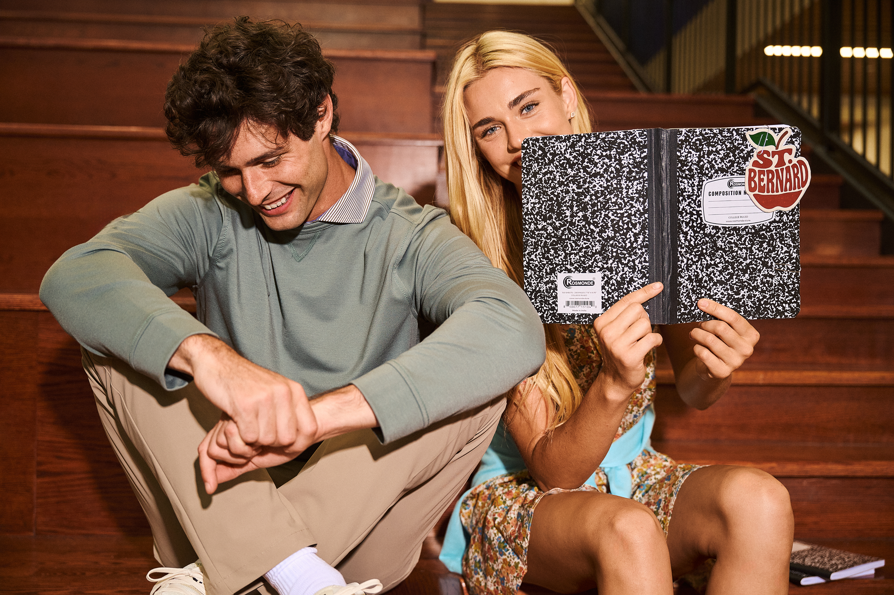 Two young adults sitting on wooden stairs, with the woman holding up a black and white marbled composition notebook.