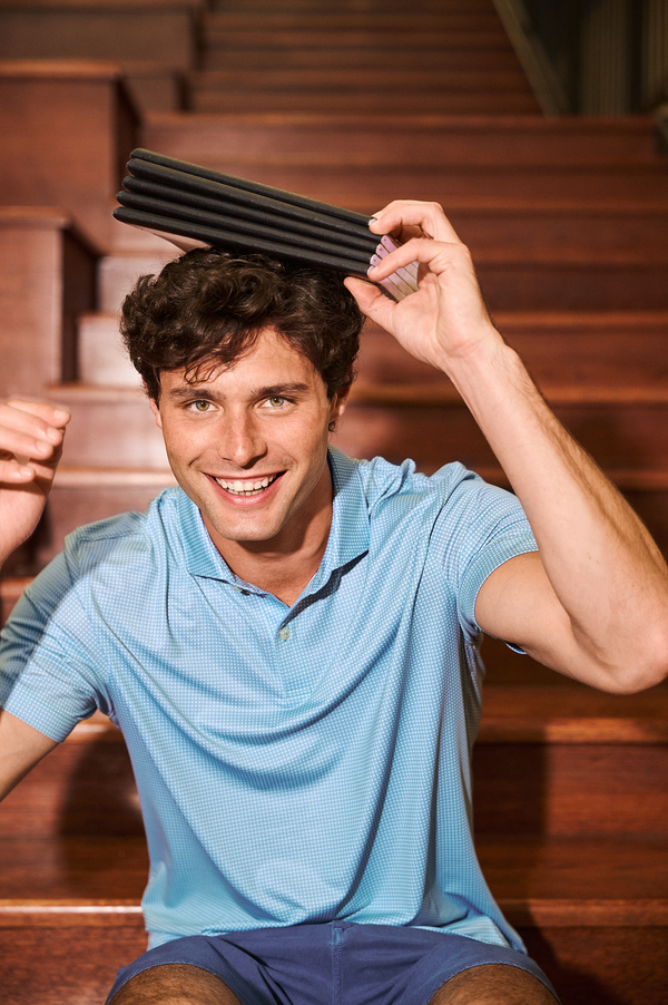 A smiling man in a light blue polo shirt and navy shorts sits on wooden stairs, balancing a couple of notebooks over his head.