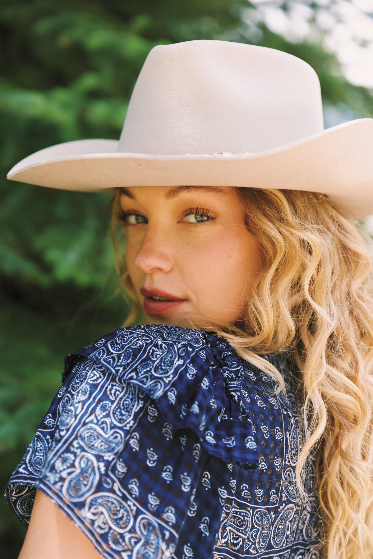 A woman outdoors wearing a light beige wide-brim cowboy hat and a blue paisley patterned top, with green foliage blurred in the background.