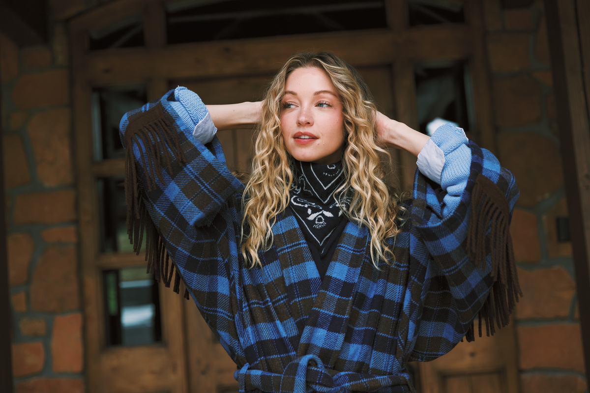 A woman with long wavy blonde hair poses in front of a rustic stone and wood building, wearing a blue and brown plaid fringed wrap jacket over a black bandana turtleneck.