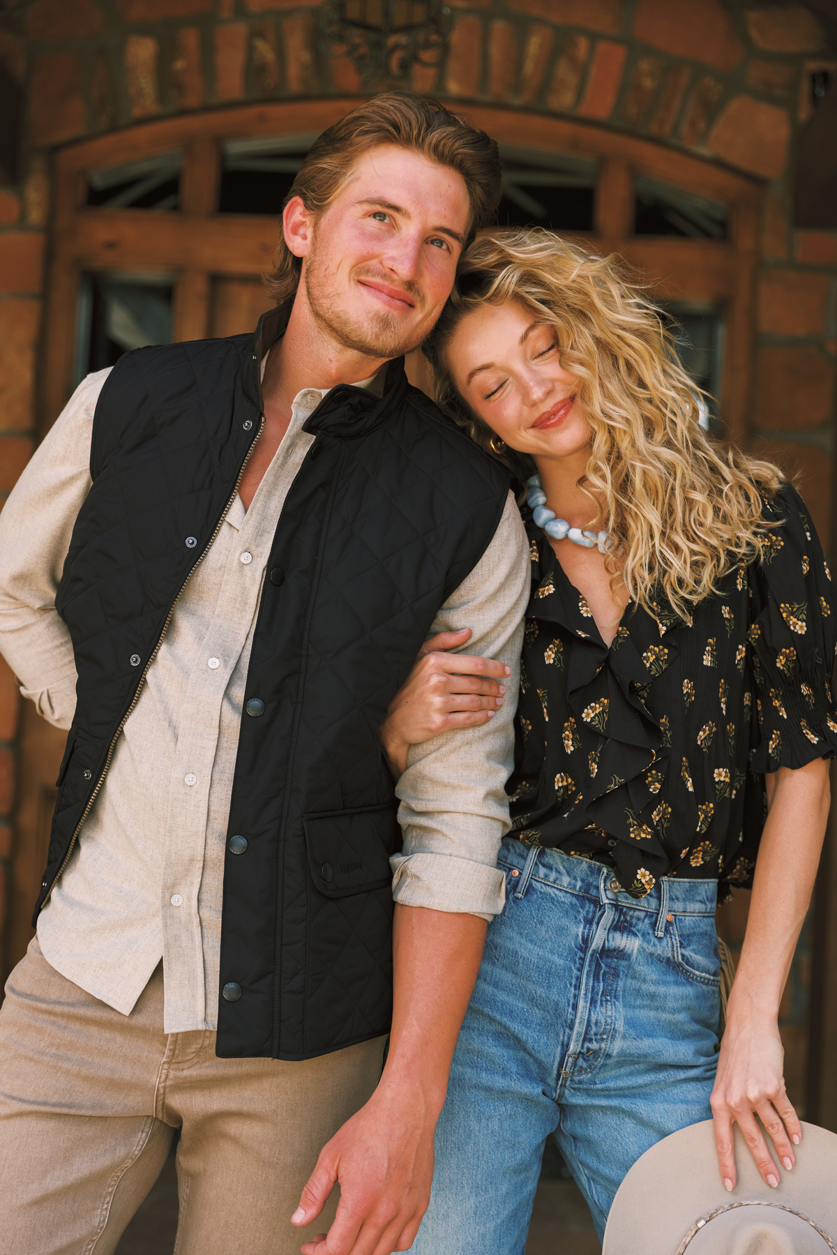 A man and a woman posing together in front of a rustic brick building. The man wears a black quilted vest over a light beige button-down shirt and khaki pants.