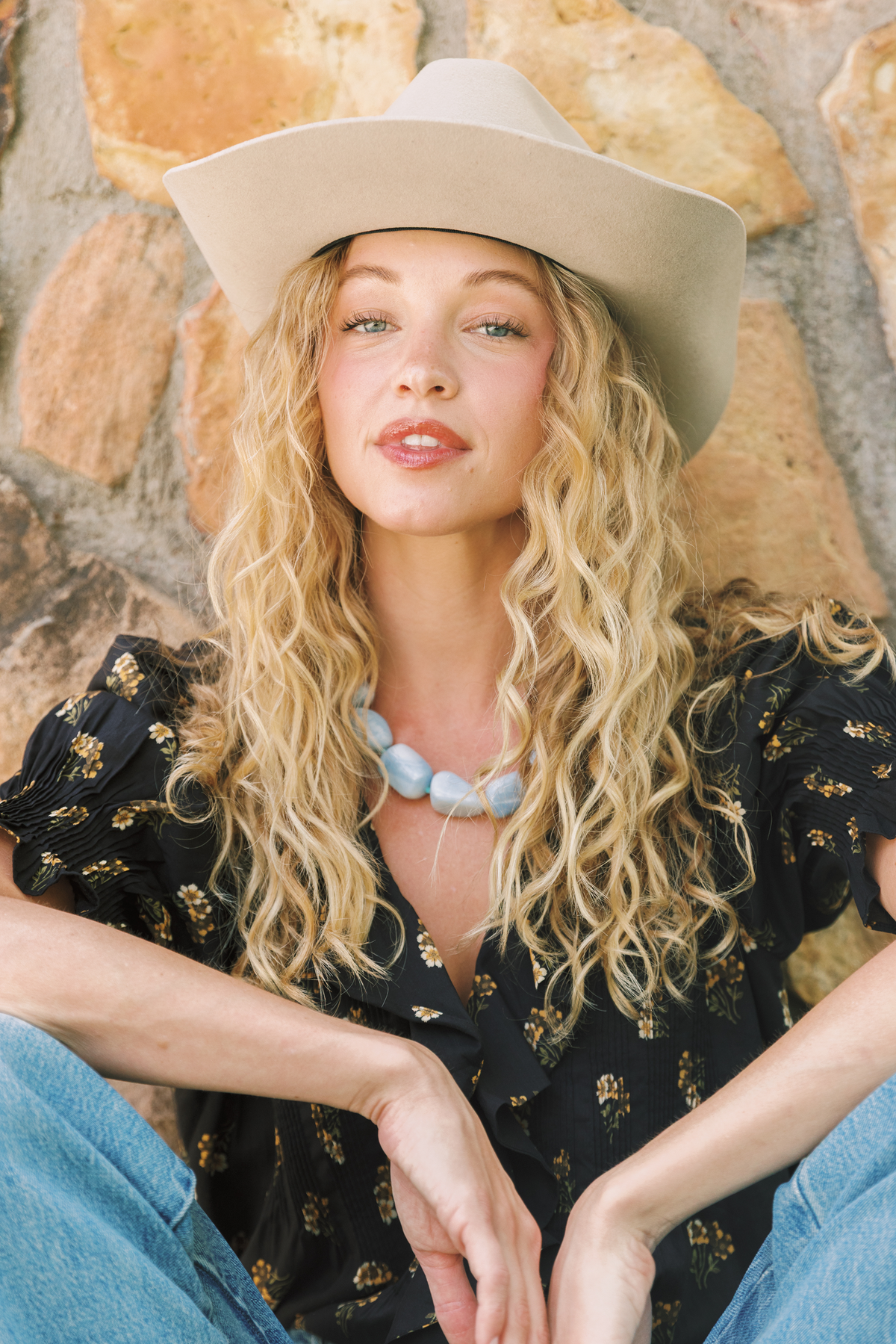 A woman with long curly blonde hair wearing a beige wide-brim cowboy hat, a black floral blouse, and blue jeans, sitting in front of a rustic stone wall.