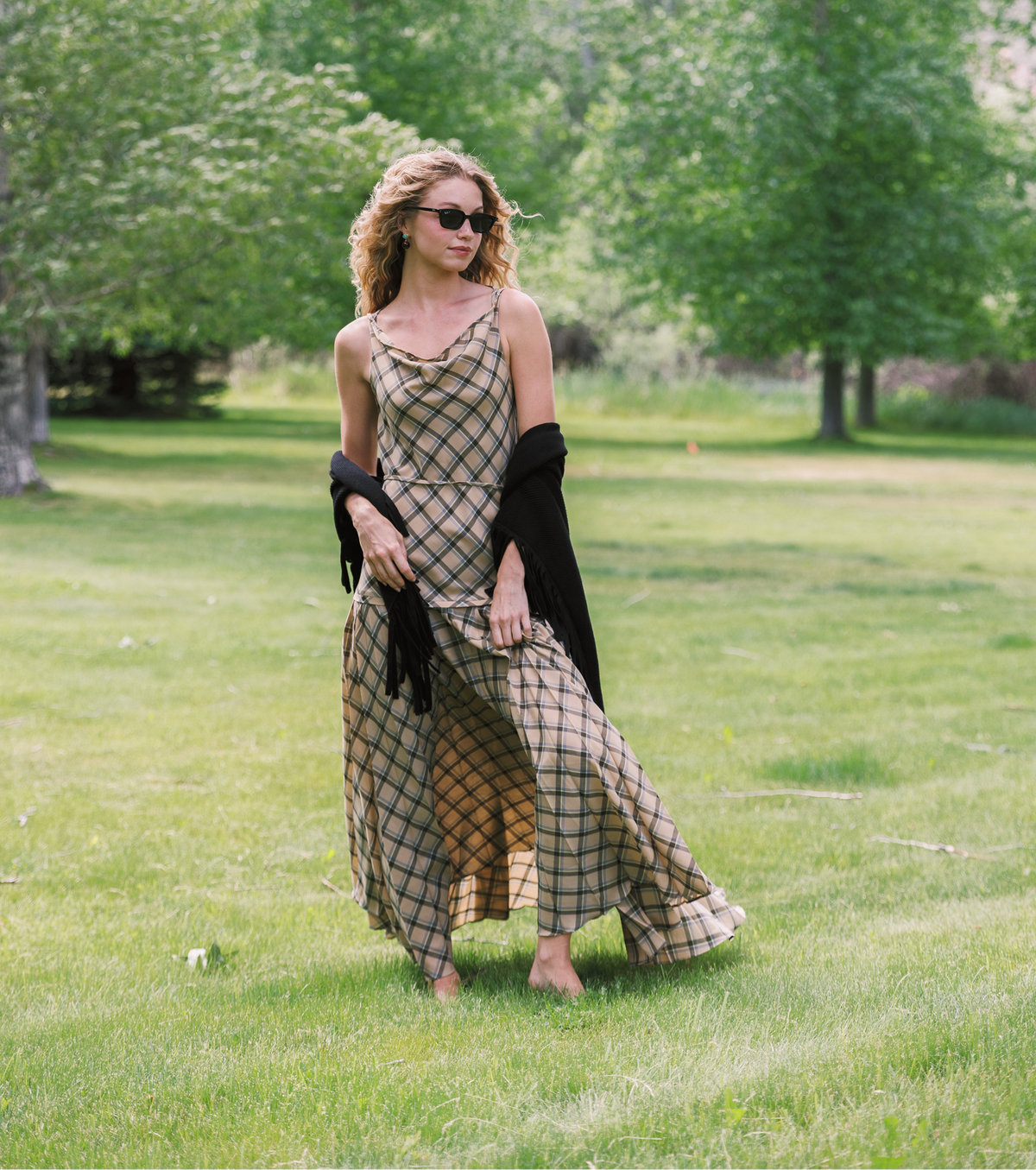 A woman stands barefoot on a grassy lawn wearing a beige and black plaid maxi dress, a black cardigan draped off her shoulders, and sunglasses, with trees in the background.