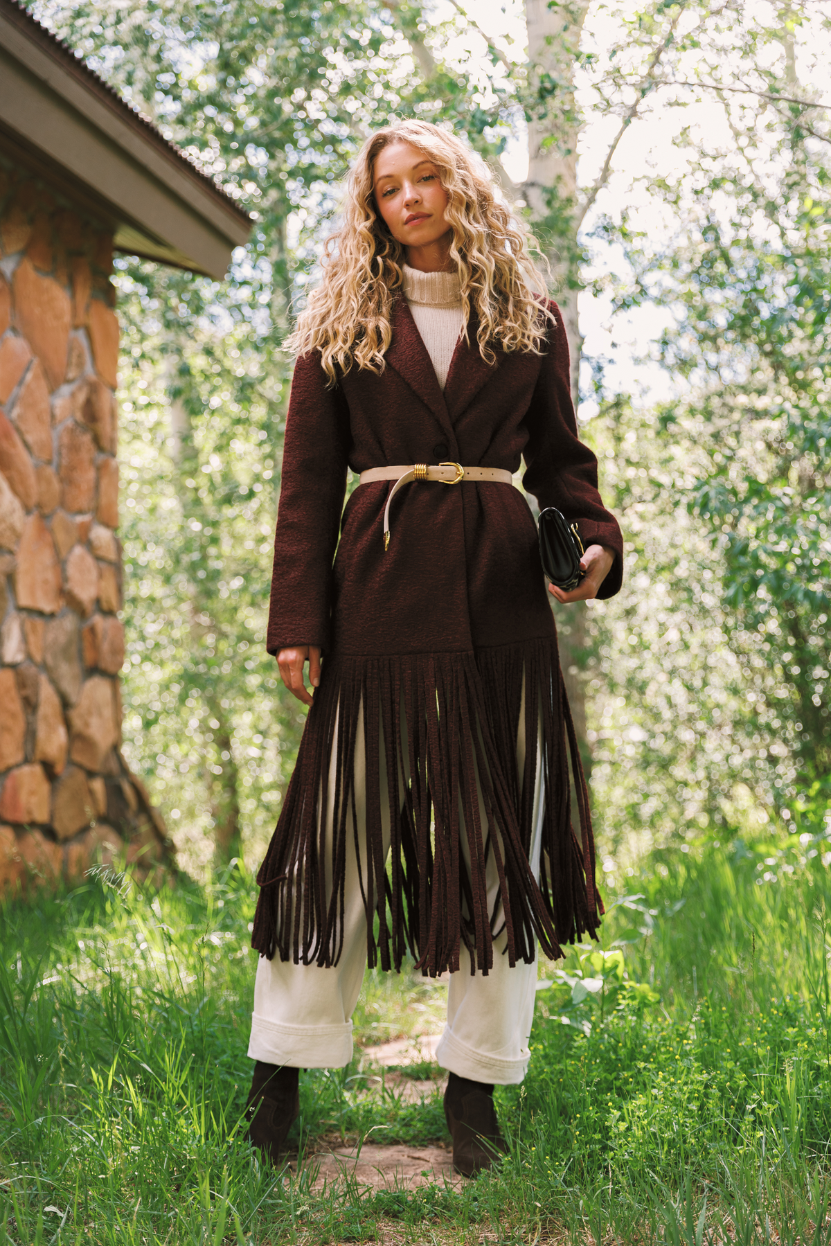 A woman stands outdoors in a grassy wooded setting wearing a dark brown long fringe coat over a cream top and white pants, with a tan belt and black boots.