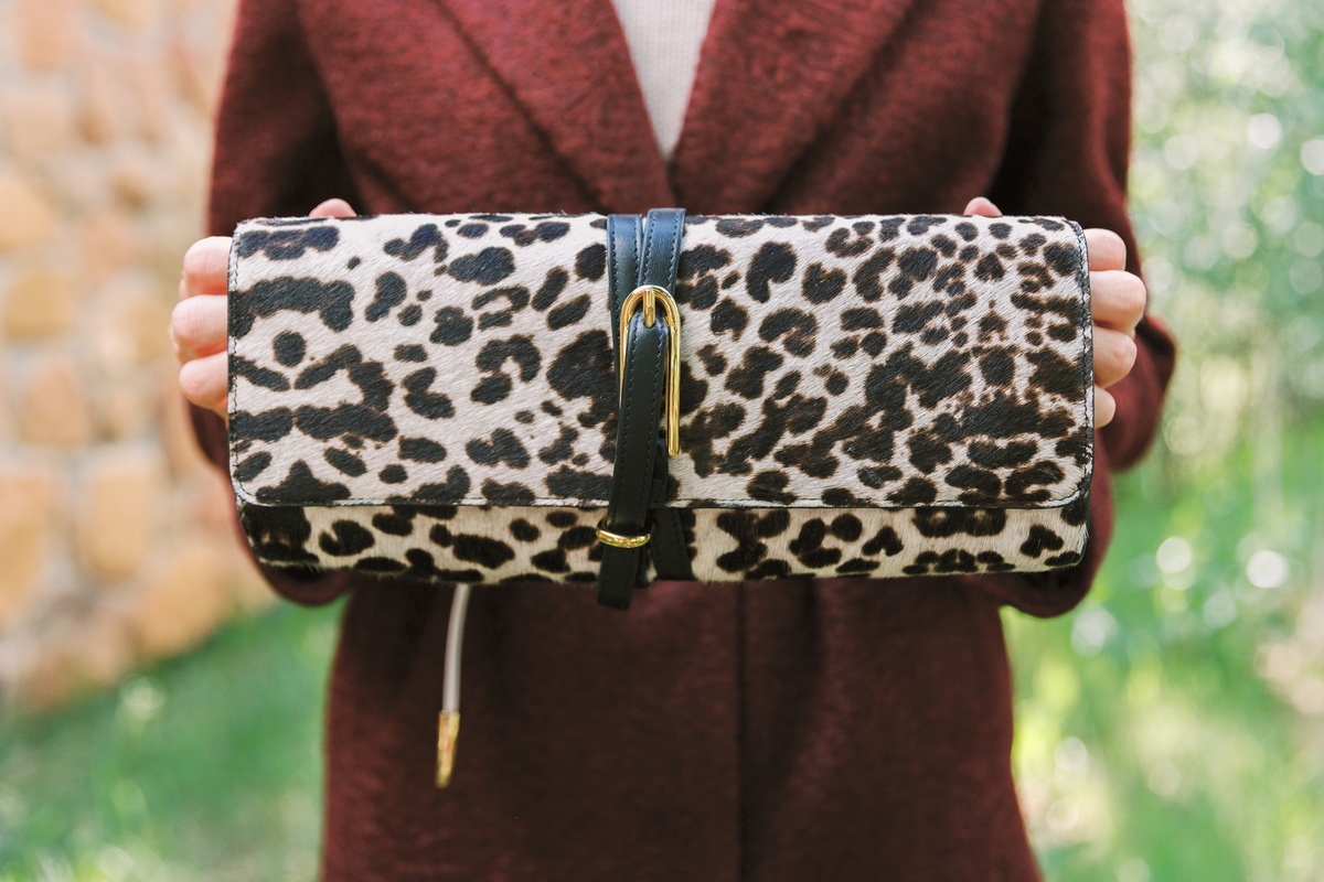 A person in a burgundy coat holds a leopard-print clutch bag with a black leather strap and gold buckle against a blurred outdoor background.
