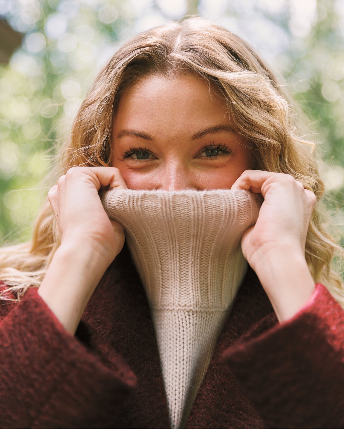 A close-up lifestyle shot of a woman outdoors, pulling a cream-colored ribbed knit turtleneck sweater up over her mouth and nose, wearing a dark brown coat. The background is a soft, out-of-focus green bokeh.