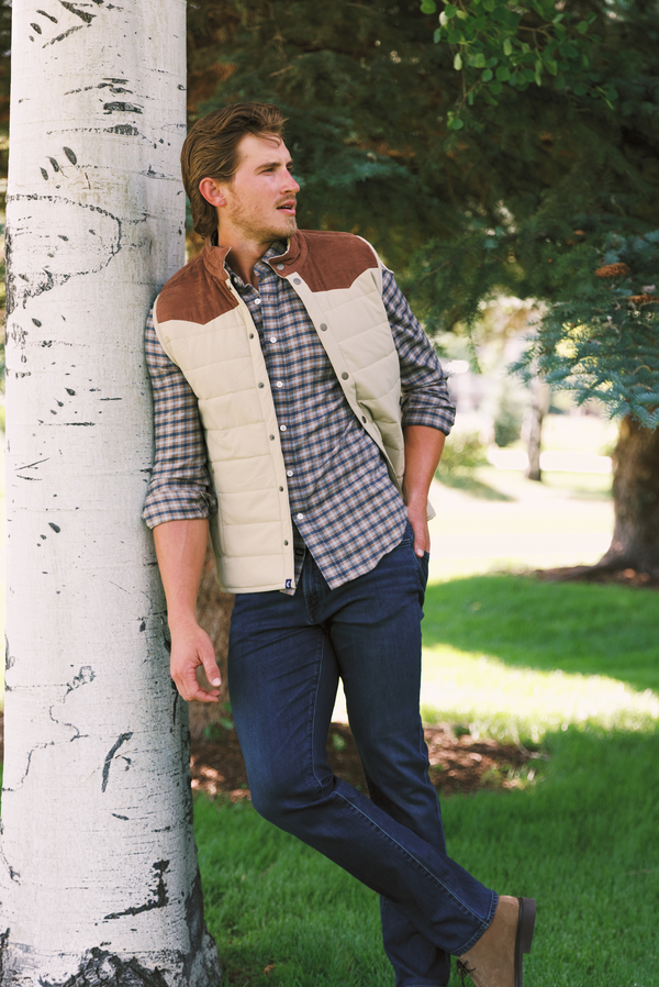 A man wearing a brown and cream quilted vest over a blue and white plaid shirt, dark jeans, and brown boots, leaning against a birch tree in a grassy park setting.
