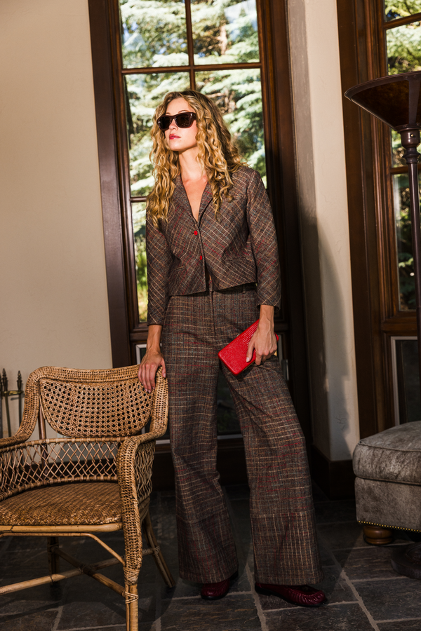 A woman stands indoors wearing a brown plaid blazer and matching wide-leg trousers, holding a bright red clutch. She is posed in front of large windows with wood trim, next to a wicker chair, with a gray upholstered ottoman visible on the right.