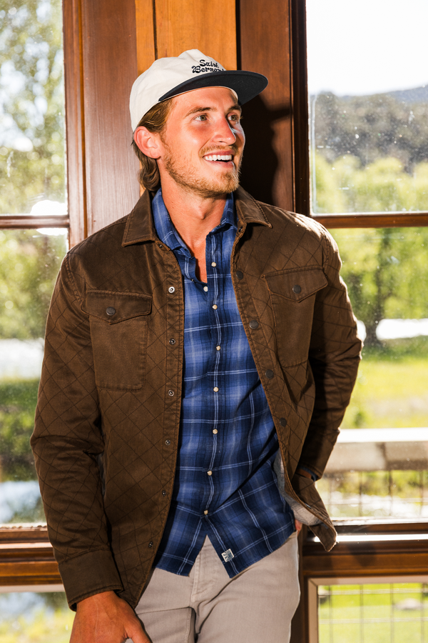 A man wearing a brown quilted overshirt, a blue plaid button-down shirt, and a white baseball cap with a black brim, standing in front of a large wooden window.