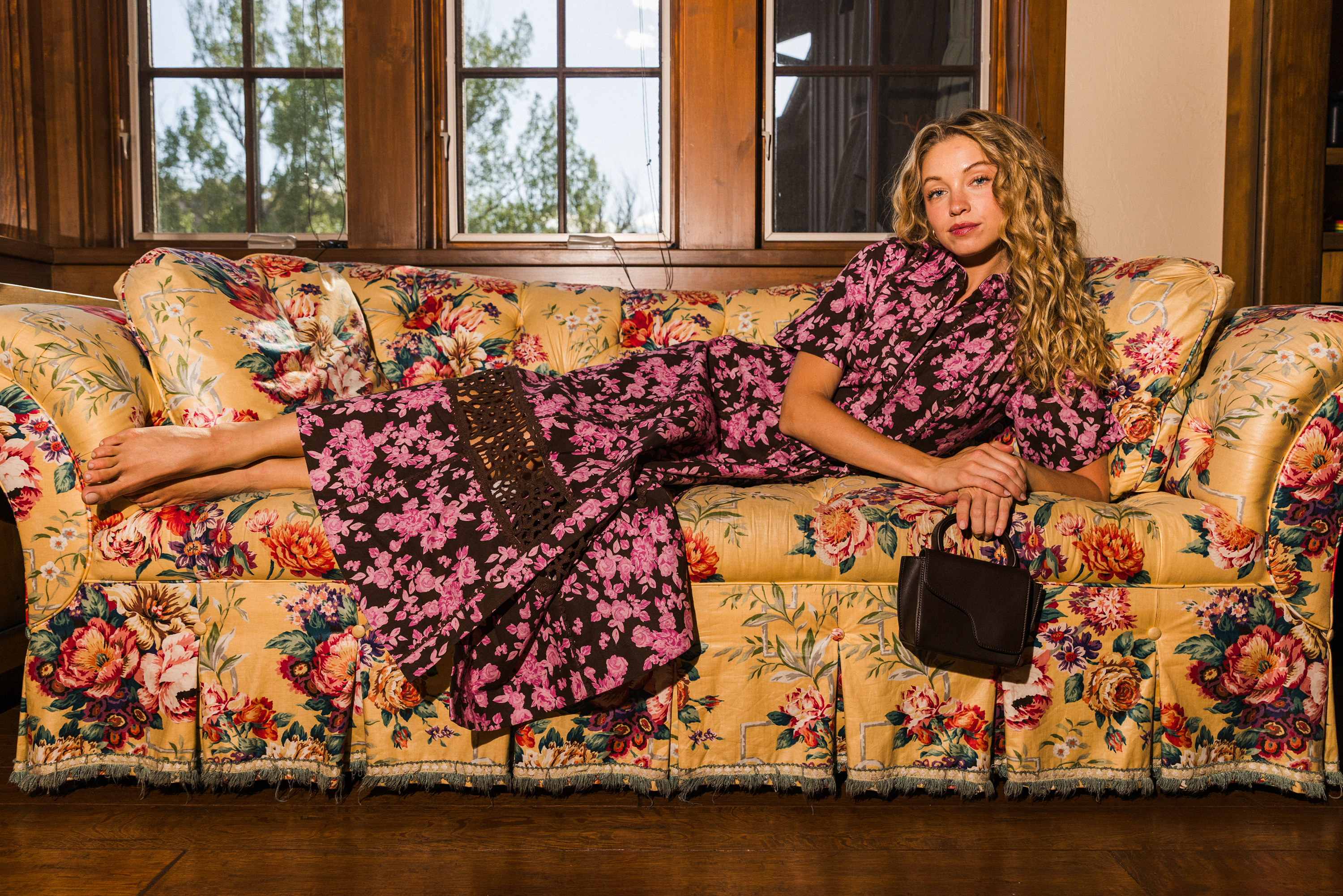 A woman reclining on a floral yellow sofa wearing a matching pink and brown patterned dress, holding a small brown handbag in a rustic indoor setting.