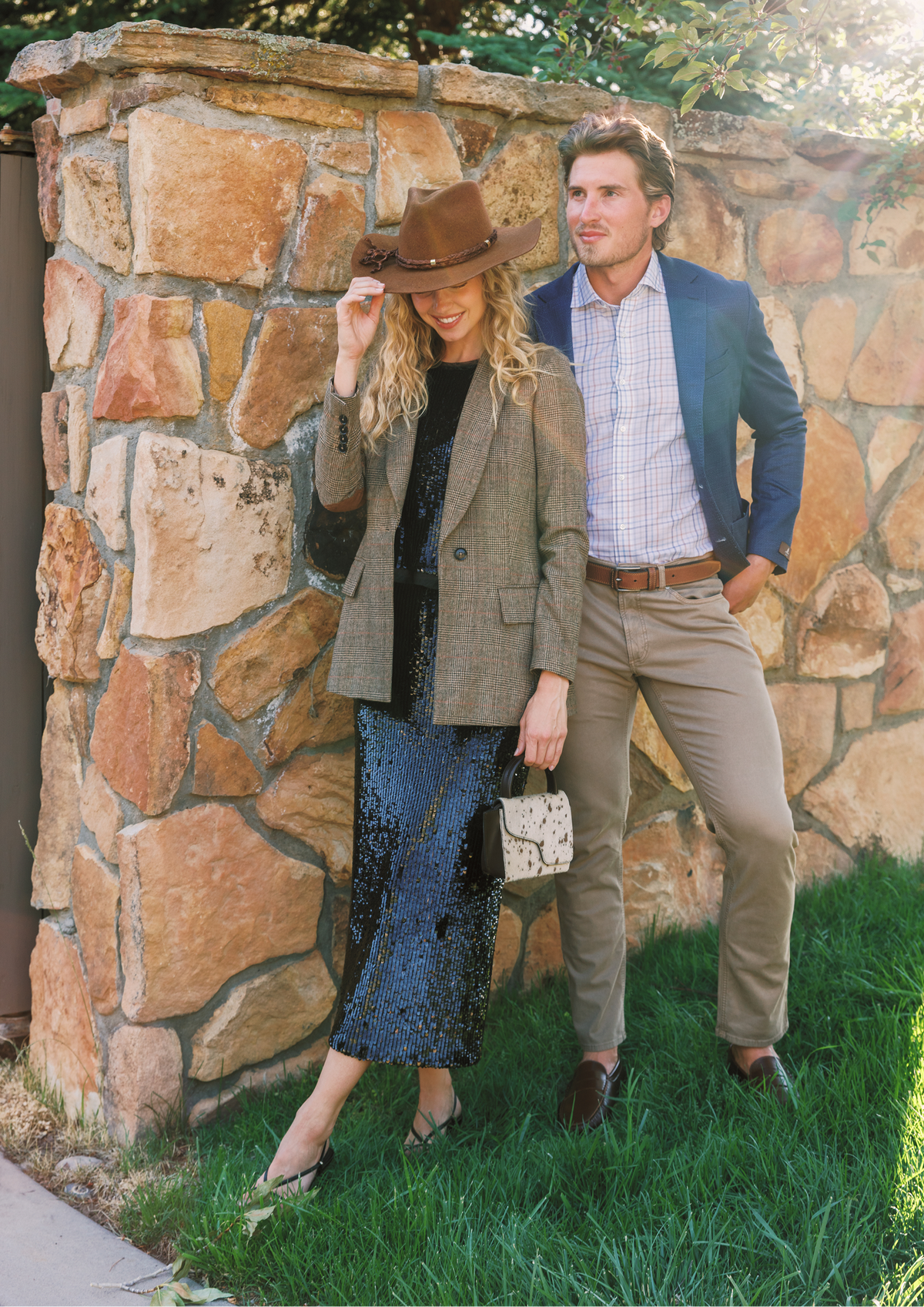 A couple posing outdoors in front of a rustic stone wall. The woman wears a brown wide-brim hat, a brown plaid blazer over a black top, and a black sequined midi skirt, holding a small patterned handbag.