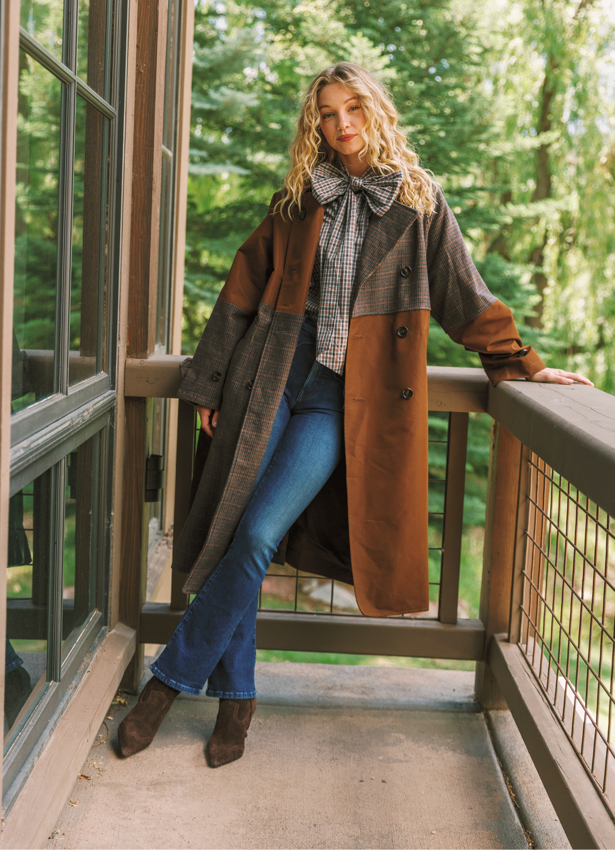 A woman leans against a wooden balcony railing wearing a long color-blocked coat in brown and gray, a plaid shirt, a large bow tie, blue jeans, and brown ankle boots, with a lush green forest background.