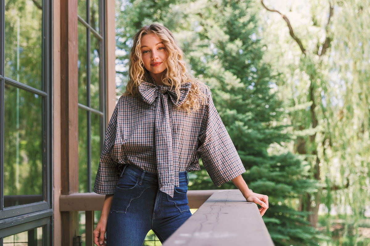 A woman stands on a balcony wearing a blue, white, and brown gingham bow blouse with wide three-quarter sleeves and dark blue jeans, with a lush green garden background.