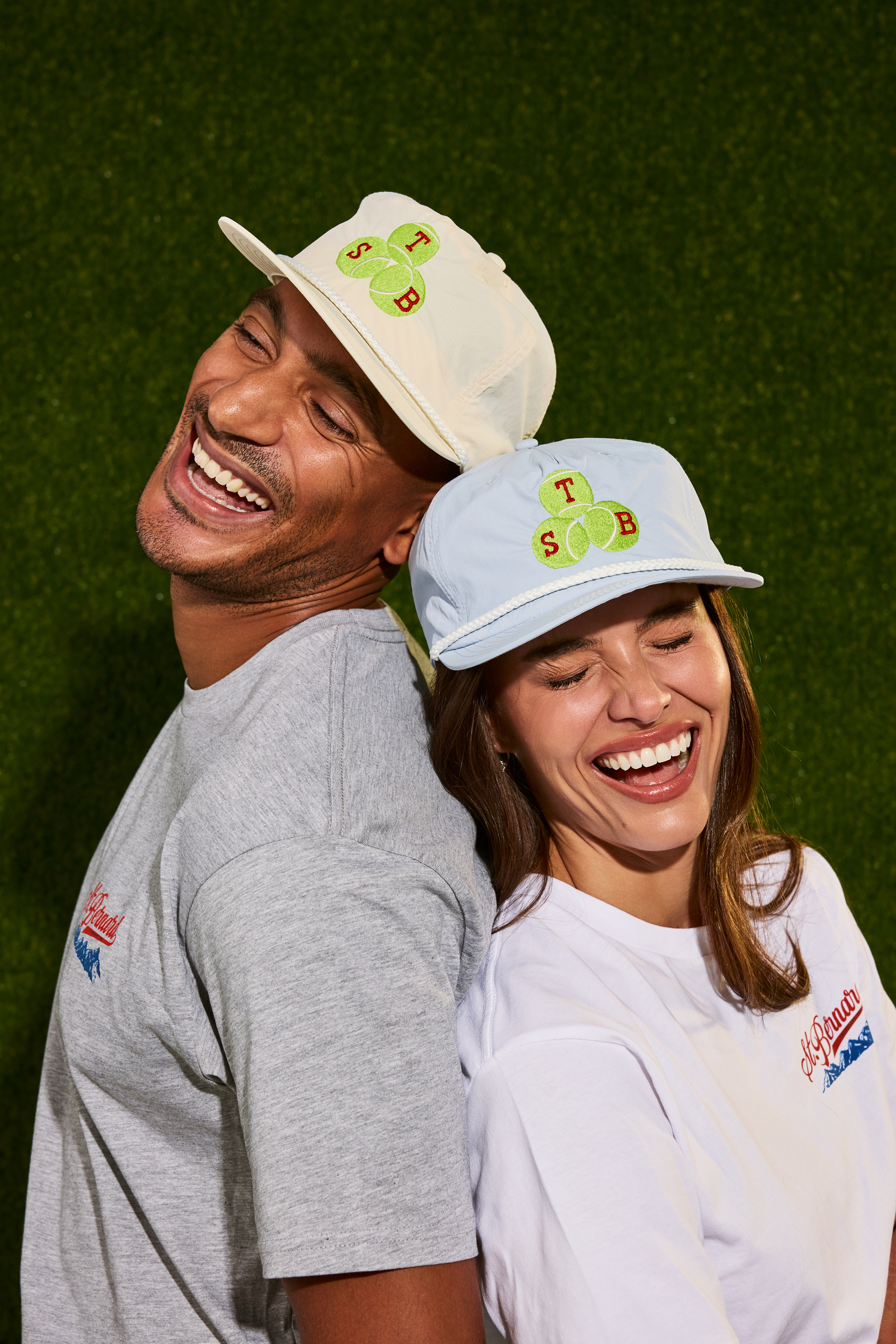A man and a woman laughing and posing together against a green artificial turf background. The man wears a cream-colored baseball cap with a tennis ball patch and a gray t-shirt, while the woman wears a light blue baseball cap with a matching teniis ball patch and a white t-shirt.