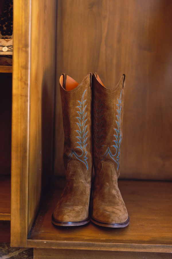 A pair of brown leather cowboy boots with turquoise embroidered floral details, standing on a wooden shelf against a wooden background.