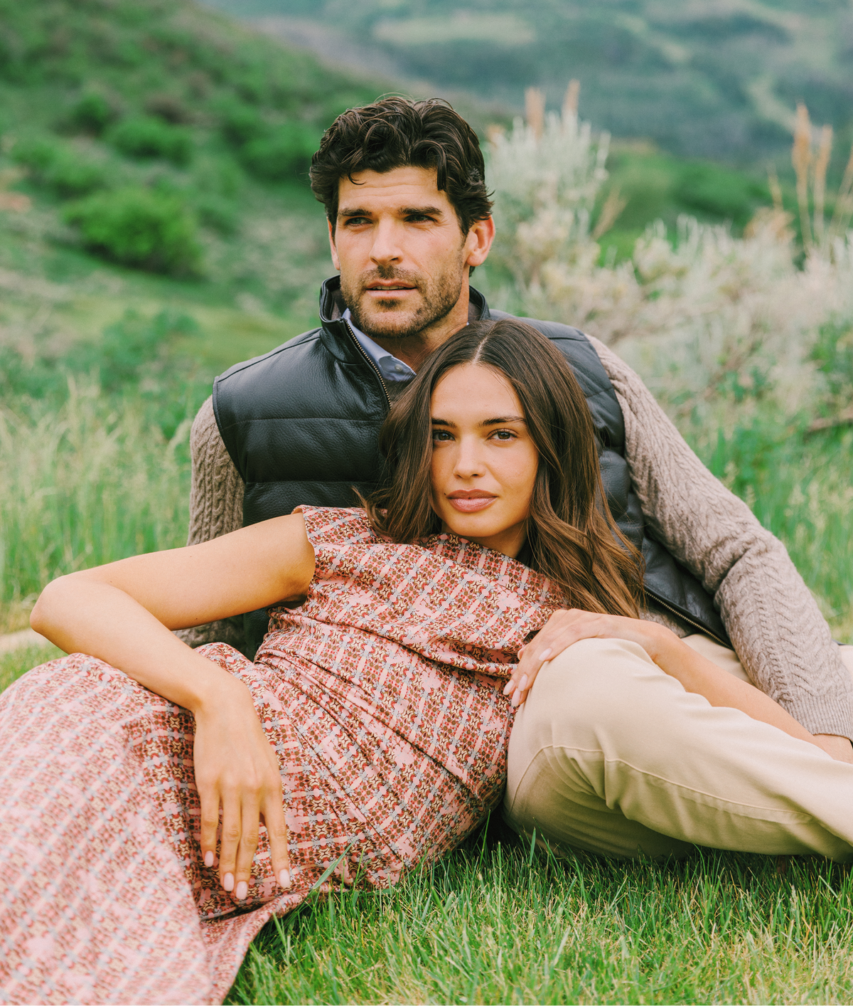 A couple posing outdoors in a grassy mountain landscape. The man wears a black puffer vest over a gray knit sweater, beige pants and the woman wears a dress.