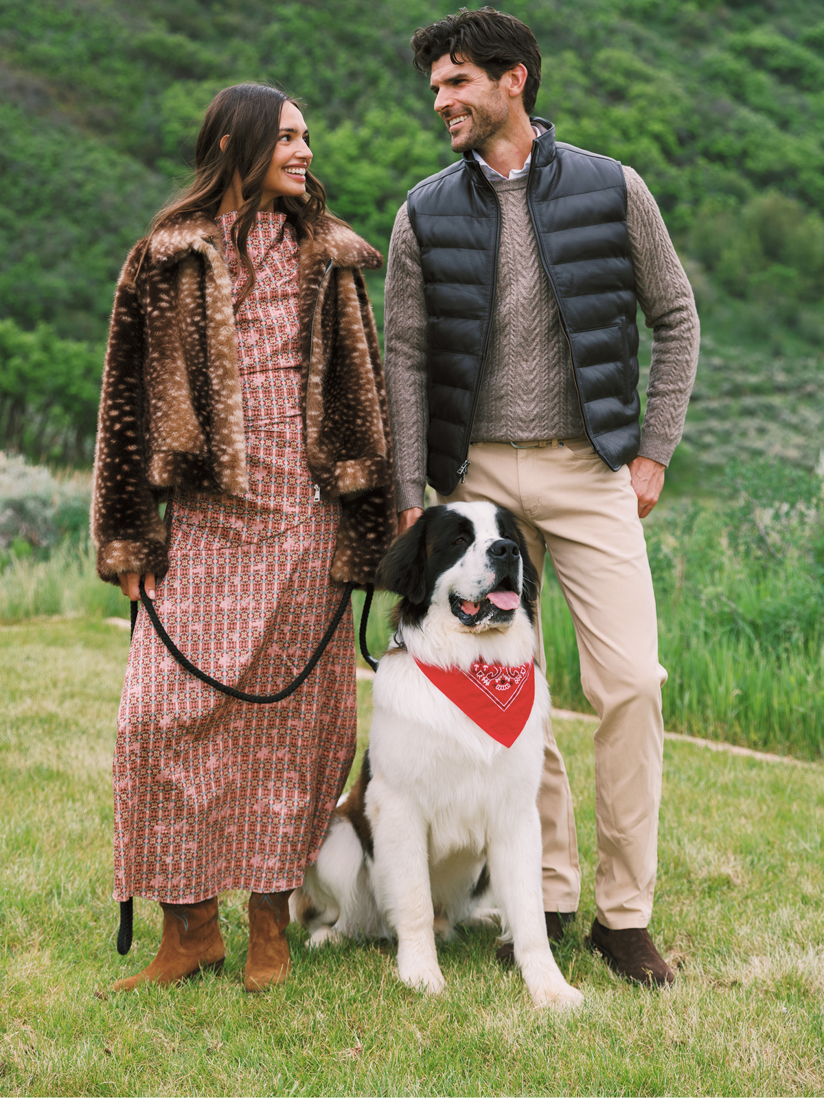 A couple and their dog pose outdoors in a grassy field with green hills in the background. The woman wears a patterned dress and a fur brown jacket, holding a black leash.