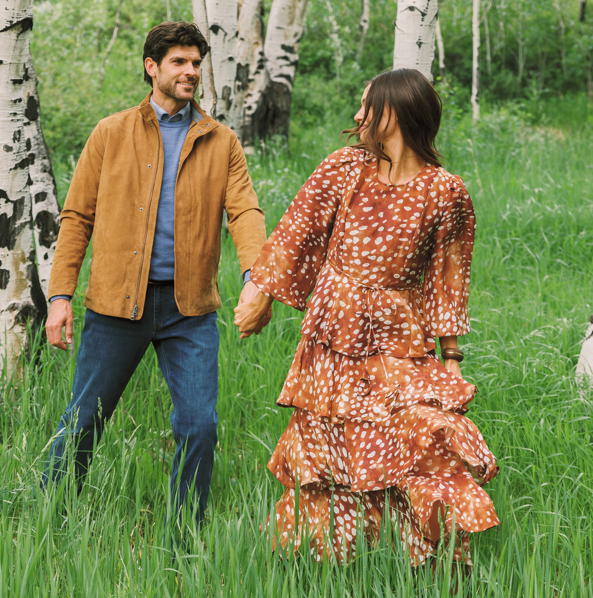 A man and a woman holding hands, walking through a grassy field with birch trees in the background. The man wears a brown suede jacket over a blue shirt and jeans, and the woman wears a rust-orange tiered maxi dress with white spots.