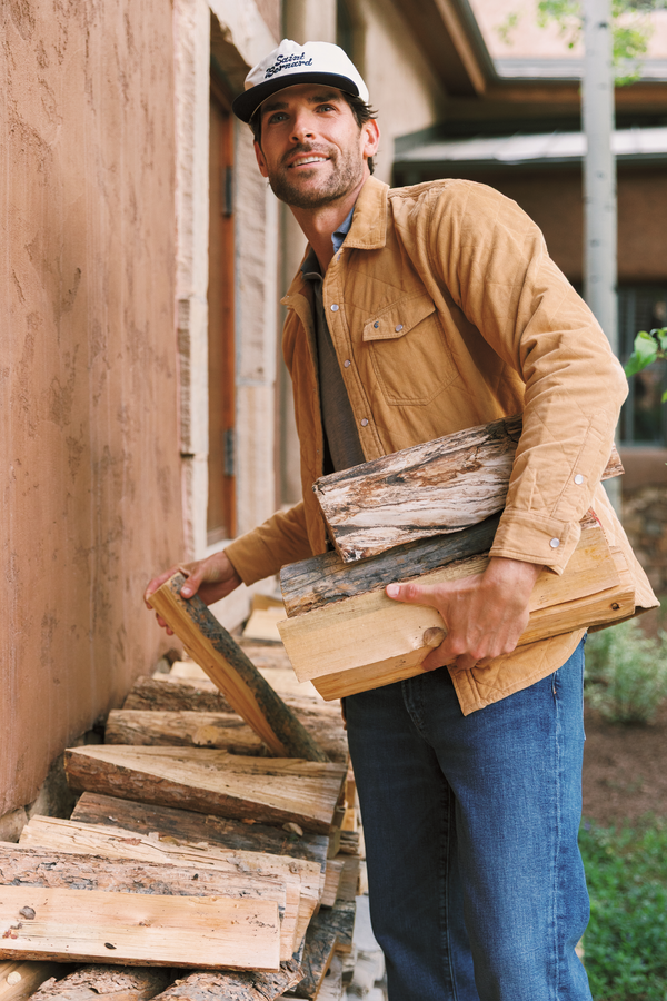 A man wearing a tan overshirt, blue jeans, and a white cap carries a stack of firewood logs next to a rustic adobe-style wall.