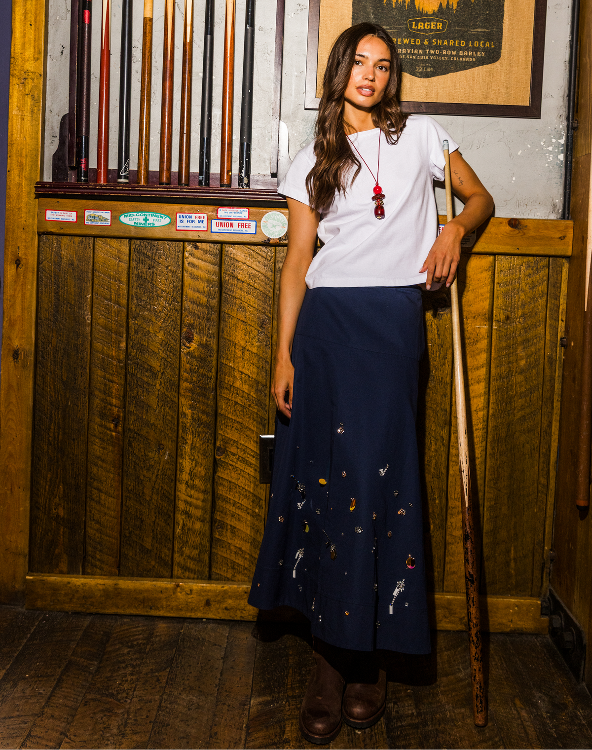 A woman stands in a rustic wood-paneled room holding a pool cue, wearing a white short-sleeve t-shirt and a dark navy blue maxi skirt with small colorful embroidered details near the hem.