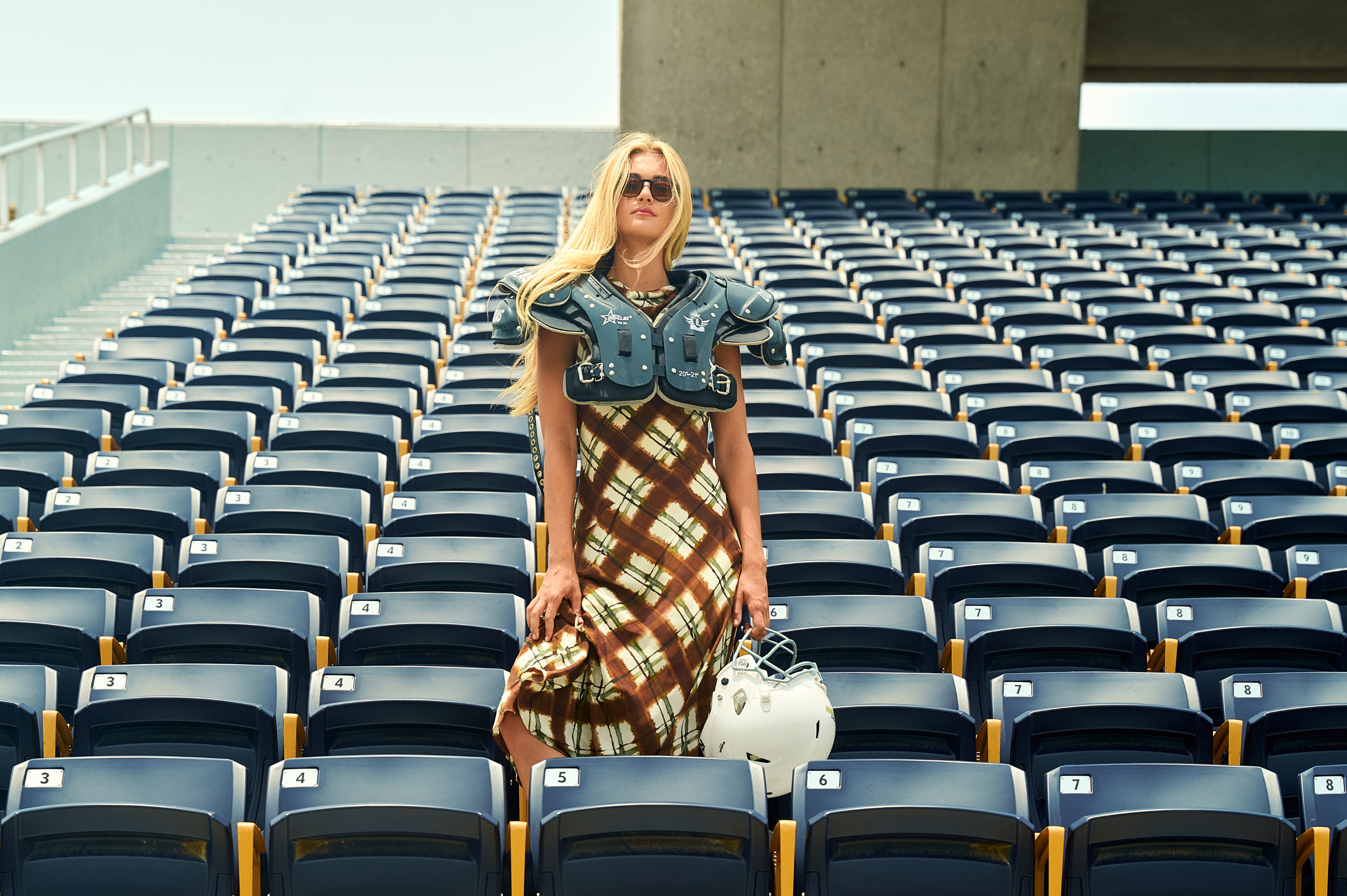 Woman in a plaid dress and blue football shoulder pads standing among stadium seats, holding a white football helmet.