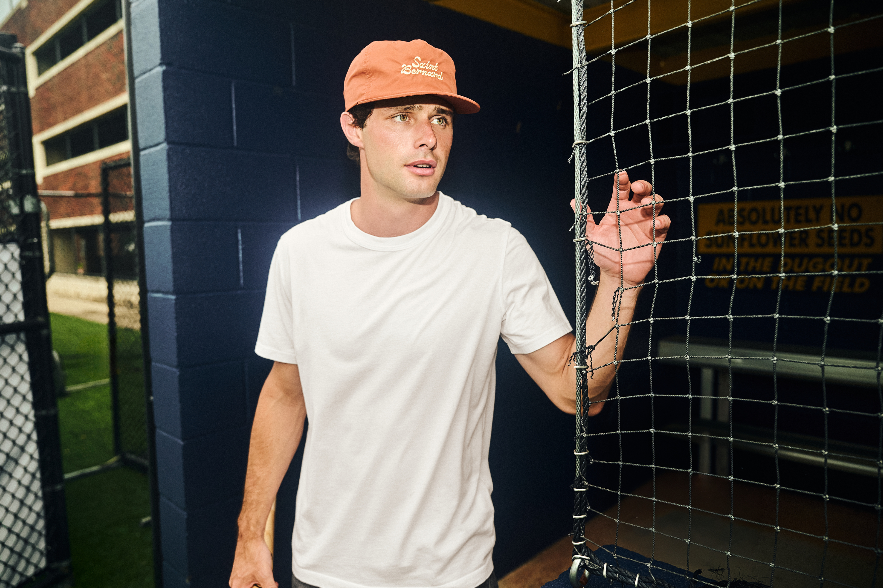 A man wearing a white t-shirt and an orange baseball cap stands next to a batting cage net in an outdoor sports facility.
