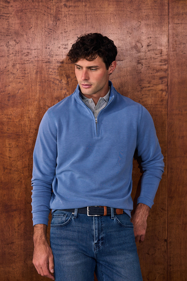 A man wearing a light blue quarter-zip sweater over a collared shirt and blue jeans, standing against a rustic brown wooden wall.