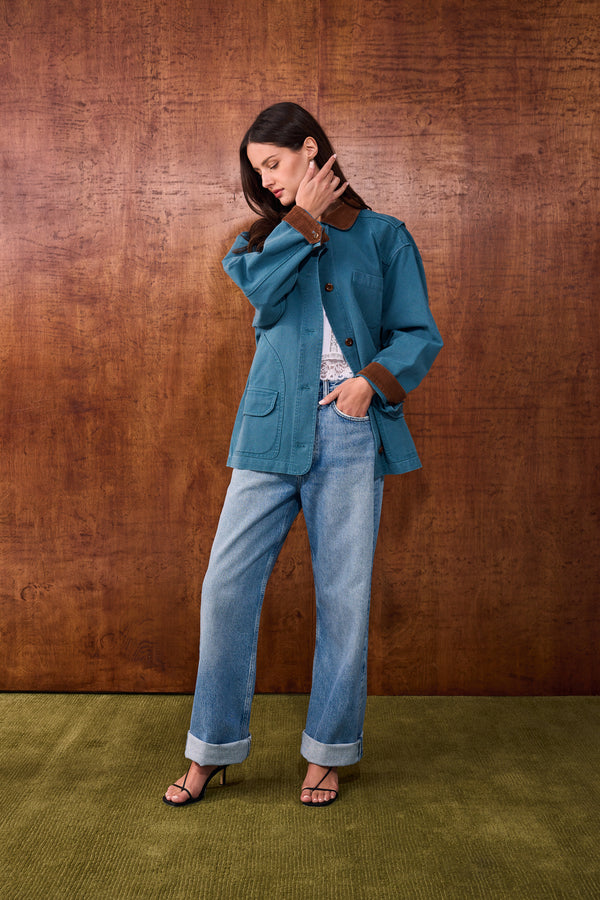A woman models a teal blue soft jacket with brown corduroy collar and cuffs, paired with light-wash blue jeans and black sandals, standing against a textured brown wall and green carpet.