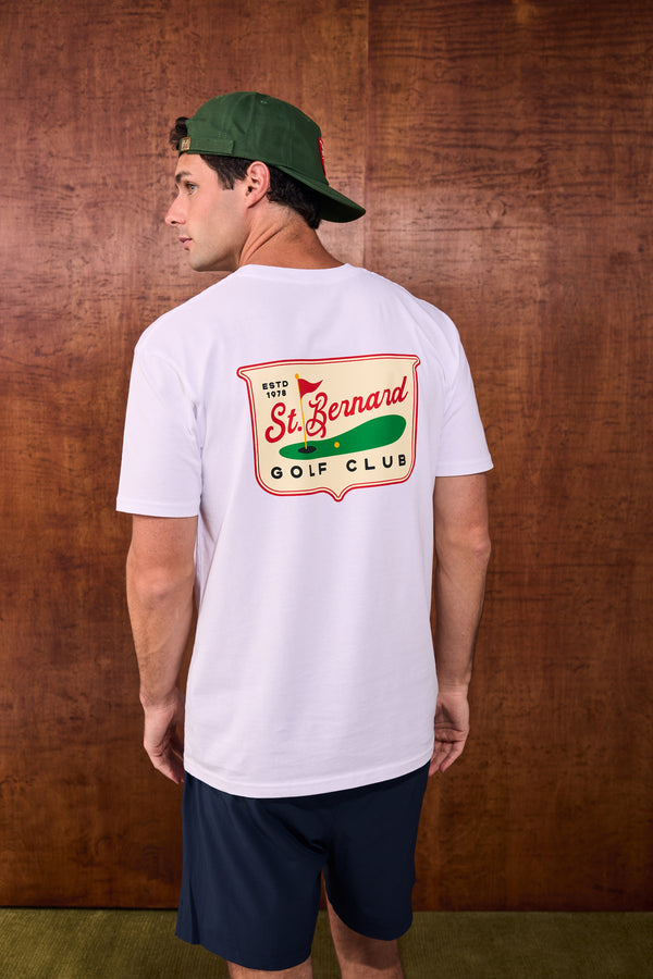 A man wearing a white t-shirt with a 'St. Bernard Golf Club' graphic on the back, navy shorts, and a green cap, standing against a brown wood-paneled wall.