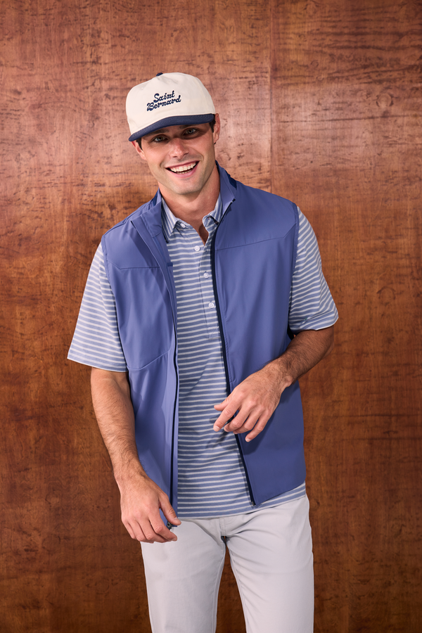 A man wearing a blue zip-up vest over a blue and white striped polo shirt, paired with a cream and navy baseball cap and light gray pants, standing against a brown wood-paneled background.