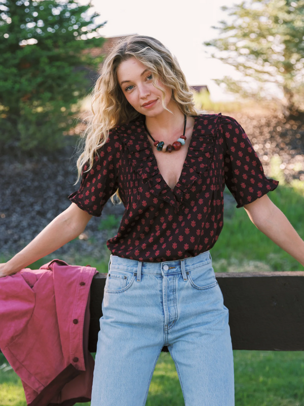 A woman wearing a dark red short-sleeve blouse with a small red floral pattern and light blue jeans, standing outdoors in front of a fence with trees in the background.