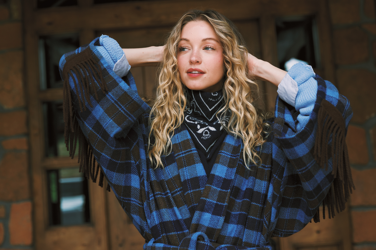 A woman with long blonde curly hair wearing a blue and brown plaid fringe wrap jacket over a navy bandana, posing with her hands behind her head against a rustic brick and wood background.