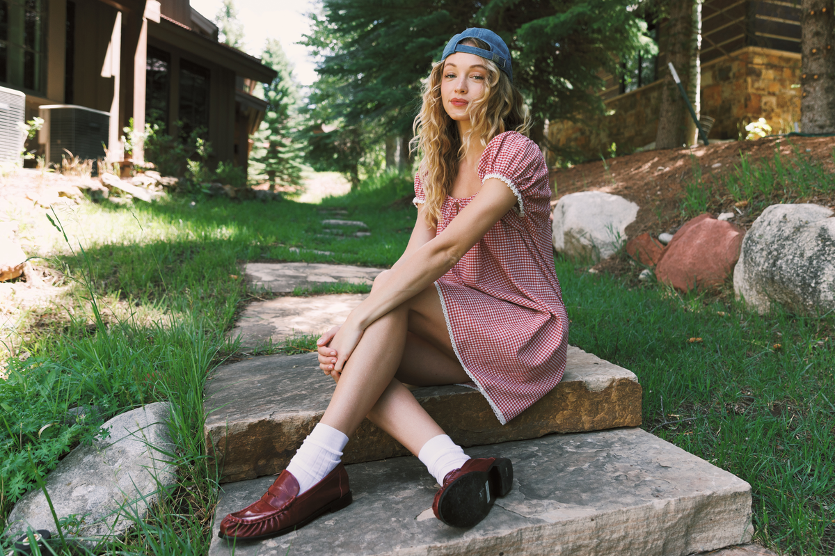 A woman sits on stone steps outdoors wearing a red gingham dress, white socks, dark red loafers, and a blue cap, with trees, grass, and rustic buildings in the background.
